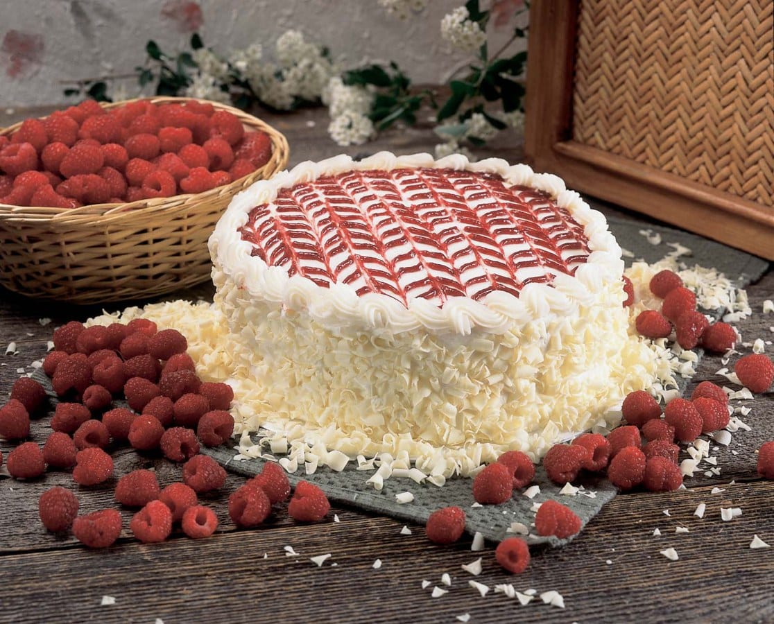 A whole white chocolate curl cake with a raspberry-glazed lattice top on a wooden surface, surrounded by fresh raspberries